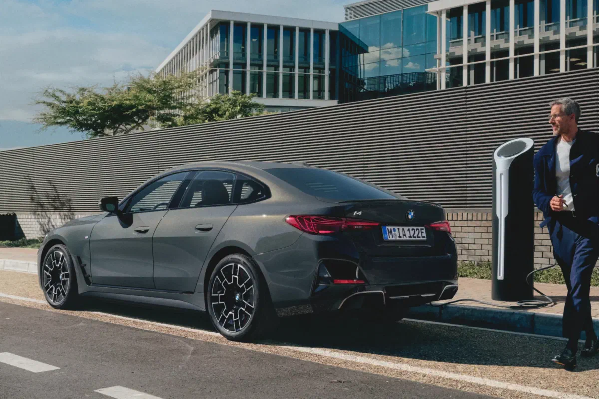 Grijze BMW i4 schuin van achteren aan een laadpaal langs de straat, met een man die naar de paal loopt.