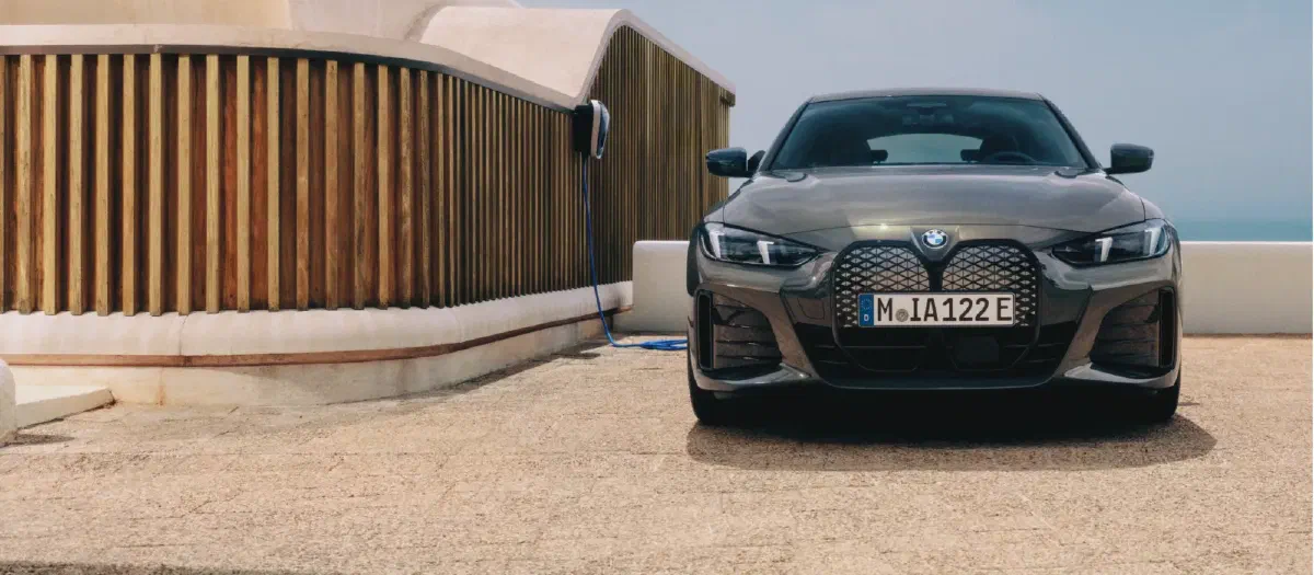 Grijze BMW i4 recht van voren op een terras aan zee, naast een wallbox aan een houten wand.