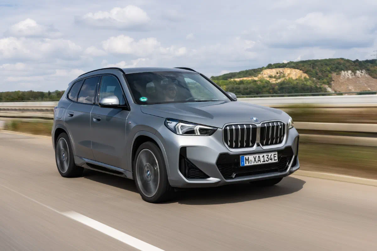 Zilvergrijze BMW iX1 met M-sportpakket rijdt over een weg, schuin van voren gezien, met een beboste heuvel en bewolkte lucht op de achtergrond.