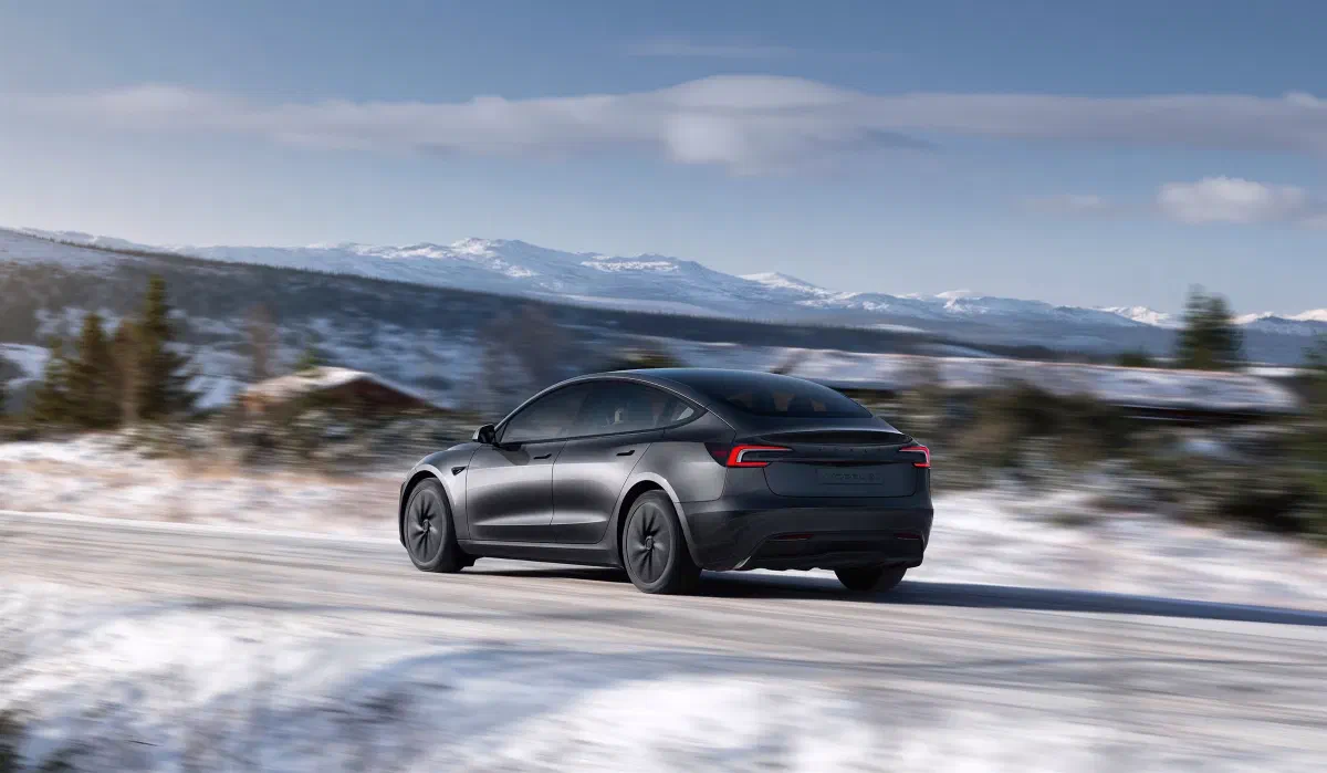 Donkergrijze Tesla Model 3 schuin van achteren rijdend over een besneeuwde weg, met besneeuwde bergen op de achtergrond.