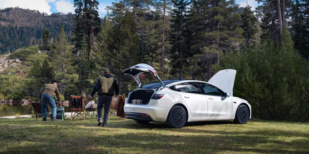 Witte Tesla Model 3 op een grasveld bij een meer in een bos, met de kofferklep en de voorklep open; twee mannen zetten campingstoelen neer.
