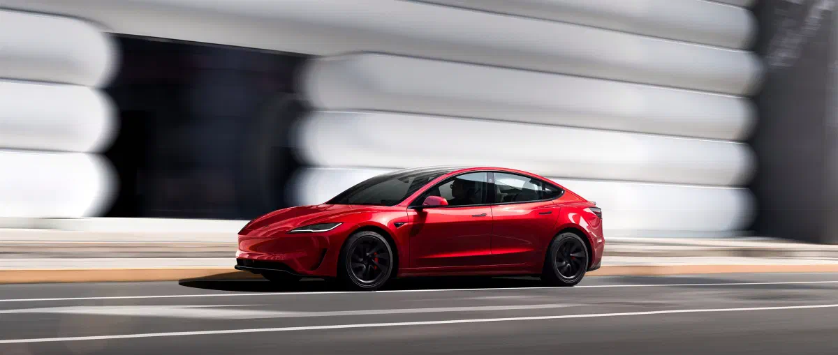 Rode Tesla Model 3 rijdend langs een wit gebouw, van opzij gezien.