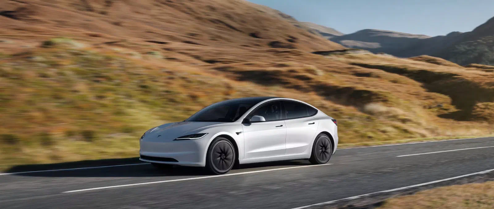 Witte Tesla Model 3 rijdend op een weg tussen bruine, glooiende heuvels.